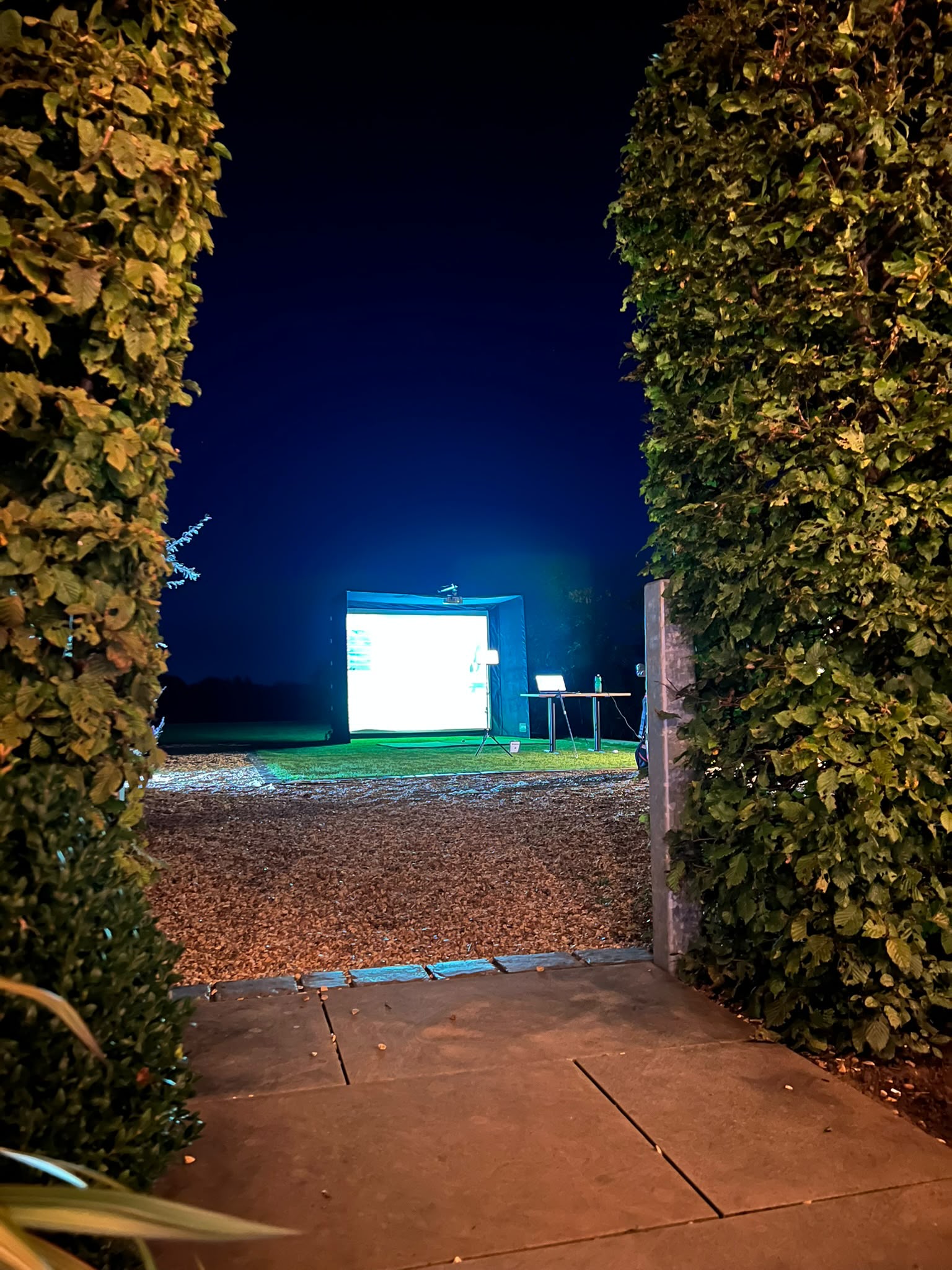 Evening golf simulator setup seen through a garden hedge